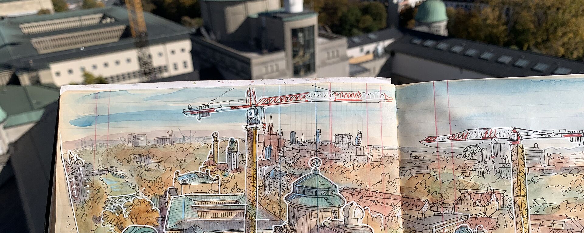 Eine Hand hält ein Skizzenbuch mit der Zeichnung der Gebäudeanlage des Deutschen Museums. Im Hintergrund sieht man das Gebäude.
