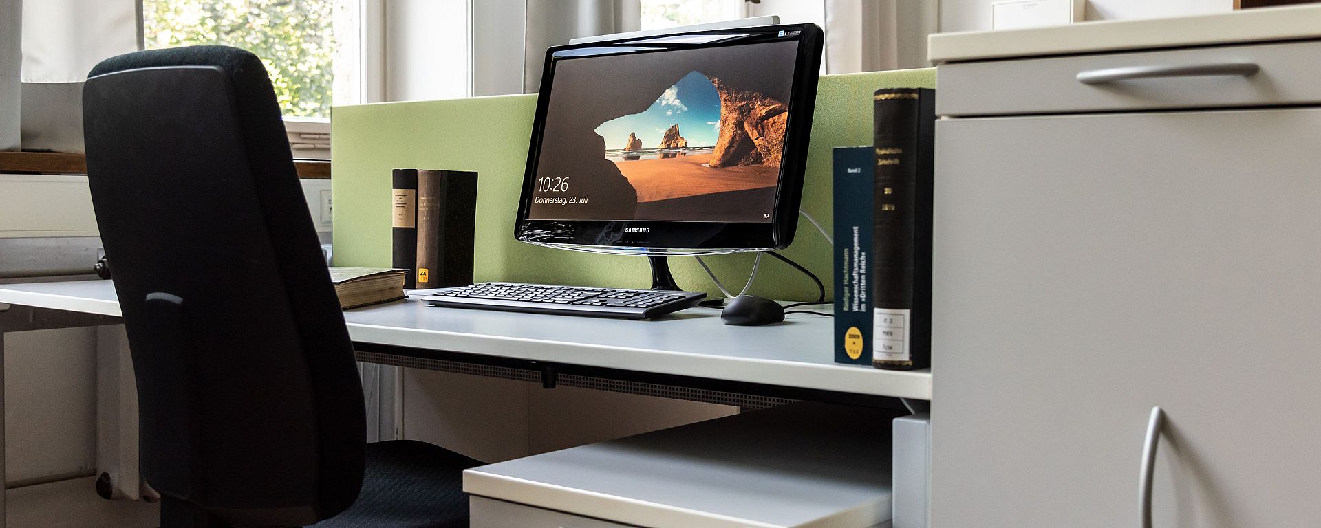 Arbeitsplatz eines Gastwissenschaftlers. Ein PC-Bildschirm und einige Bücher stehen auf dem Tisch.